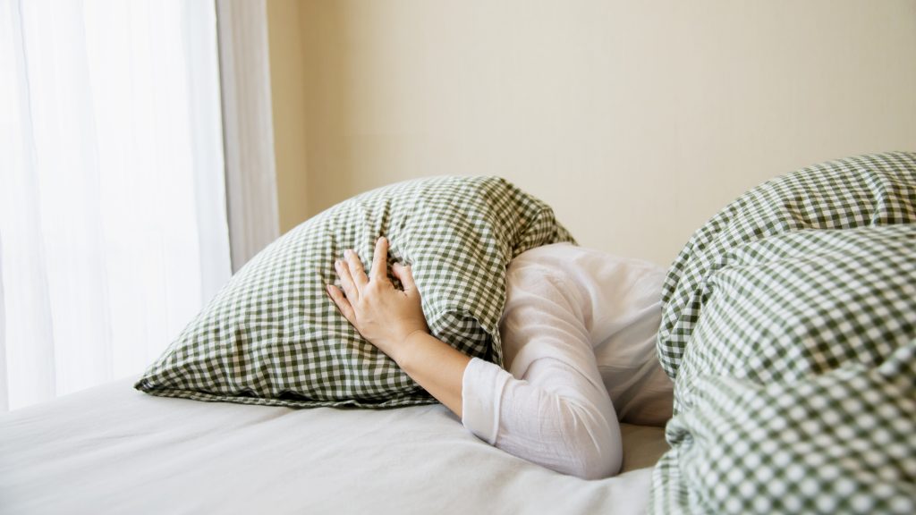 chronic insomnia patient covering her head with pillow while lying on a bed