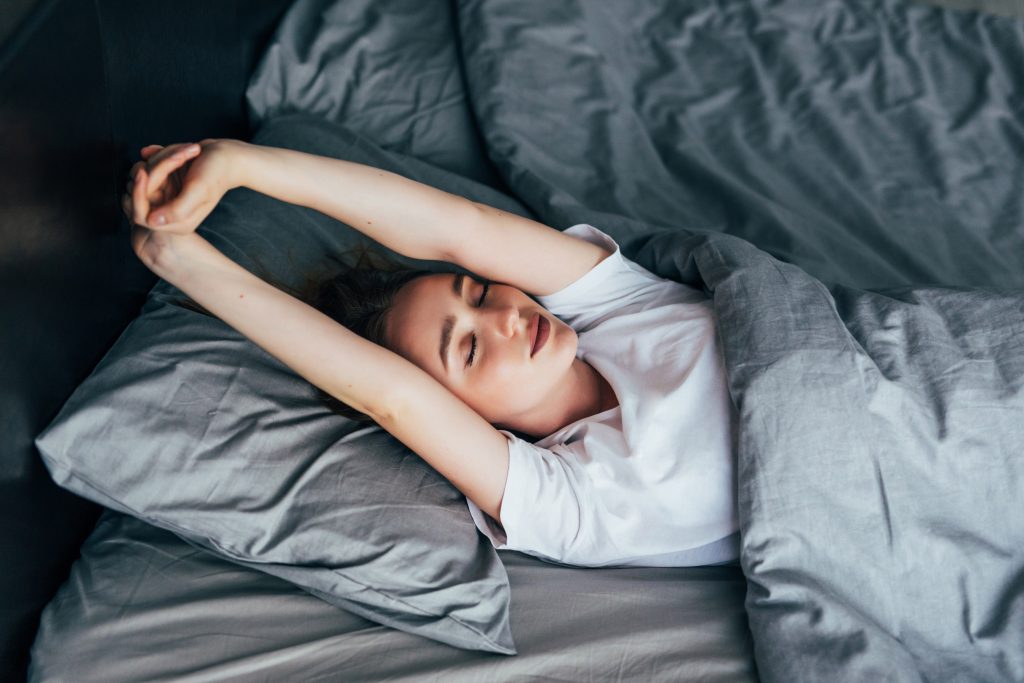 woman in a white tshirt stretching her arms over her head while lying in a bed