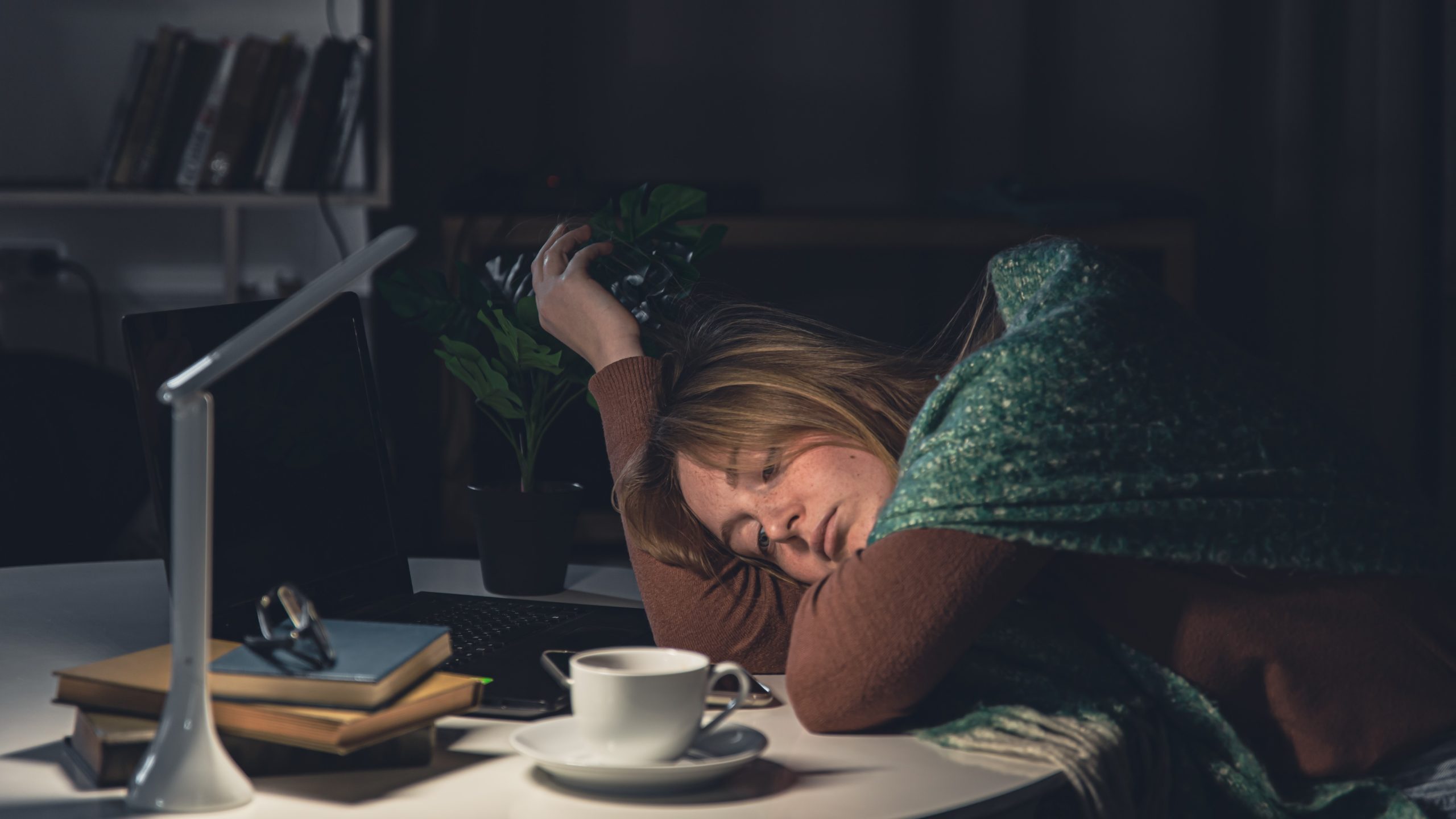 exhausted woman leaning her head on a desk late at night next to a laptop and coffee cup