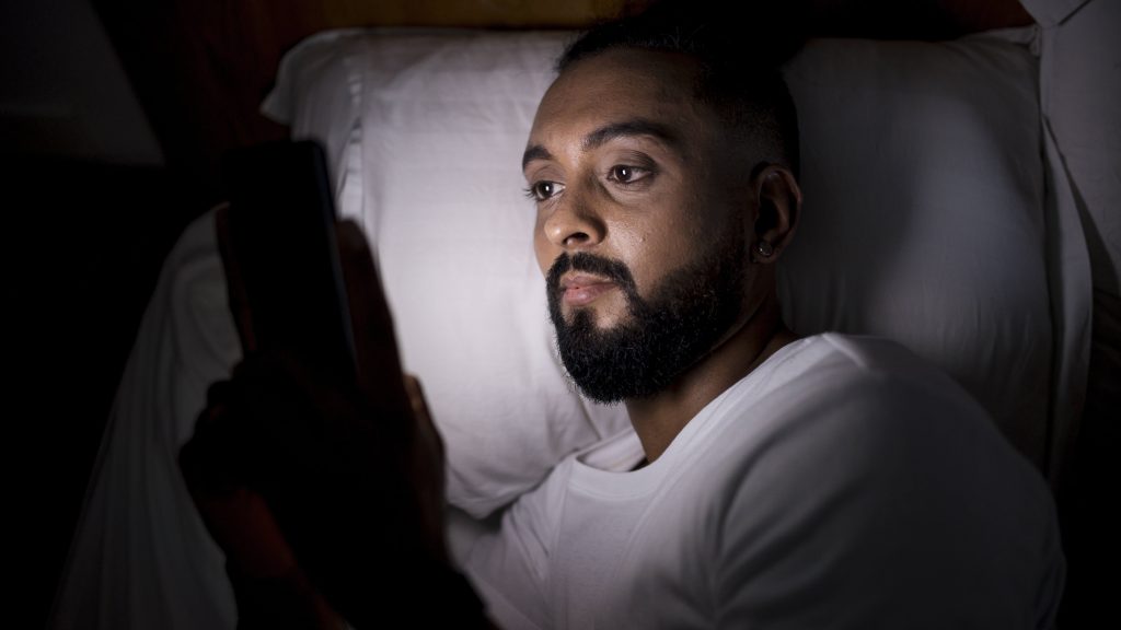 a man looking at his smartphone in bed late at night causing sleep deprivation